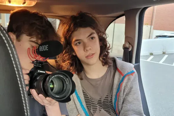 Two teenagers from In Focus Teens sit in a car’s back seat—one films with a Canon camera and Rode mic, while the other glances at the lens. The cozy car interior frames them, with a parking lot just outside the window.