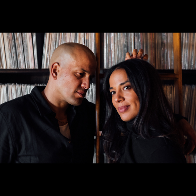 Two people stand before shelves of vinyl records. The bald person on the left looks down; the other, with long dark hair, smiles at the camera. Both wear dark clothes, capturing a vibe reminiscent of JOHNNYSWIM’s Georgica Pond era.