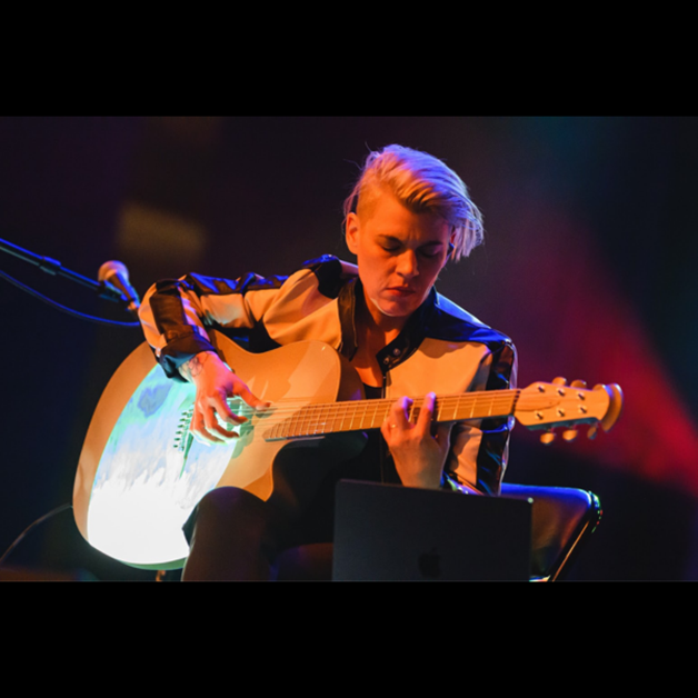 A person with short, blonde hair sits on stage playing an acoustic guitar. They wear a patterned jacket and are focused on the instrument. Stage lighting in blue and red hues illuminates the scene. A microphone is visible nearby.