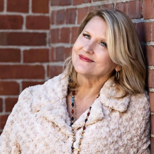 A woman with blonde hair in loose waves, resembling Medium Lauren Robbins, wears a textured coat and beaded necklace. She leans against a reddish-brown brick wall, smiling slightly as she gazes upward.