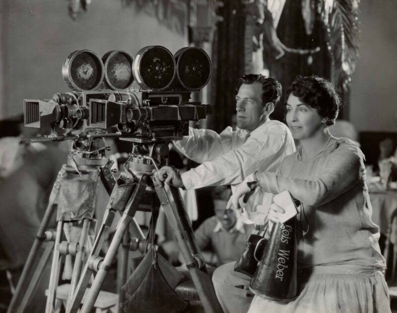 A man and a woman operate a large vintage film camera on a tripod. The woman holds a megaphone labeled “Lois Weber.” Both appear focused, and the background shows blurred figures and set decorations. The image is in black and white.