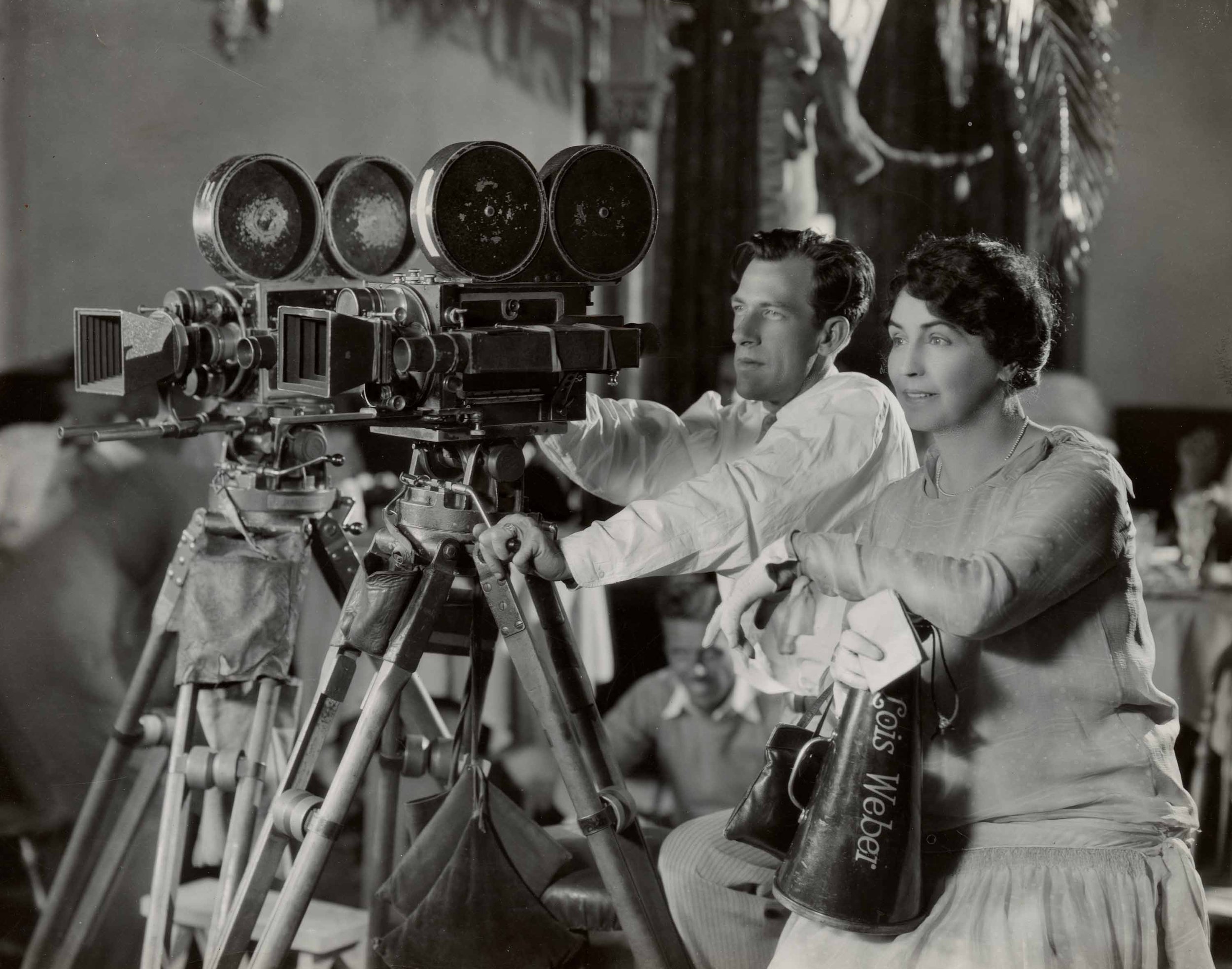 A man and a woman operate a large vintage film camera on a tripod. The woman holds a megaphone labeled “Lois Weber.” Both appear focused, and the background shows blurred figures and set decorations. The image is in black and white.