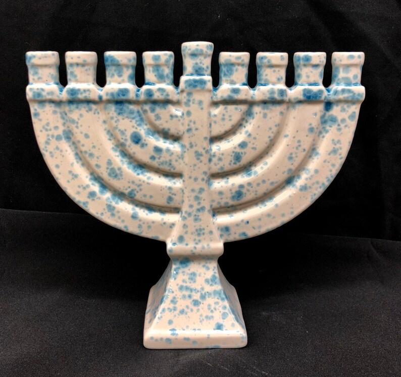 A ceramic menorah with eight candle holders and a central holder, featuring a white base with blue speckles, is displayed against a black background—perfect inspiration for your next Menorah painting or family event.