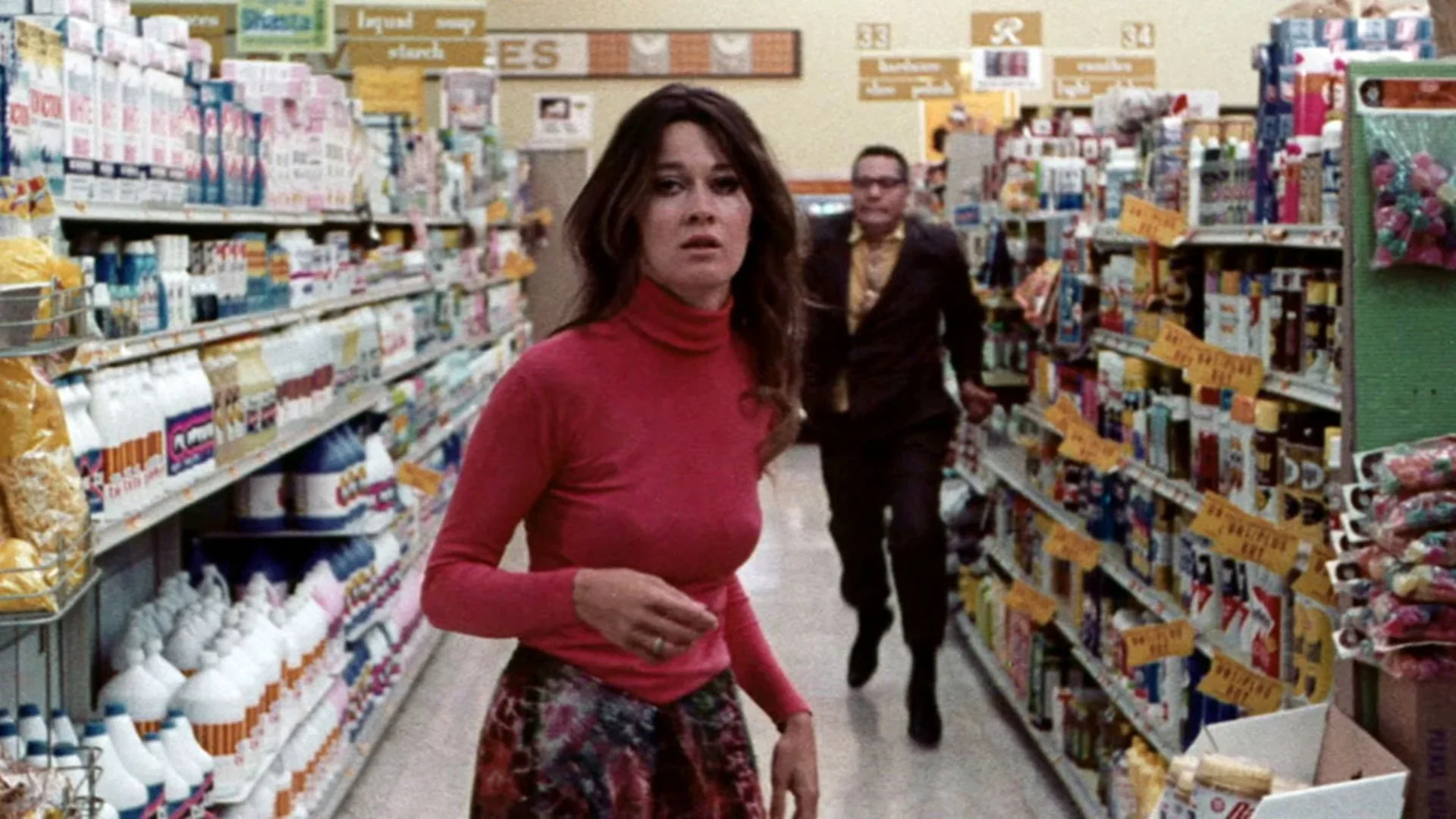 A woman in a red turtleneck and floral skirt walks quickly down a grocery store aisle, looking concerned. A man in a suit follows behind her. Shelves are stocked with various products, and yellow sale signs hang above the aisles.