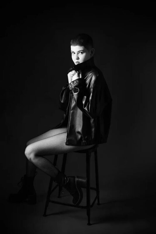 A person with short hair sits on a stool in a dark studio, wearing a leather jacket, fishnet tights, and ankle boots. They look at the camera and hold the collar of the jacket up, with dramatic lighting highlighting their face and outfit.