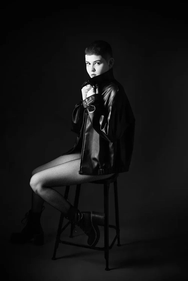 A person with short hair sits on a stool in a dark studio, wearing a leather jacket, fishnet tights, and ankle boots. They look at the camera and hold the collar of the jacket up, with dramatic lighting highlighting their face and outfit.