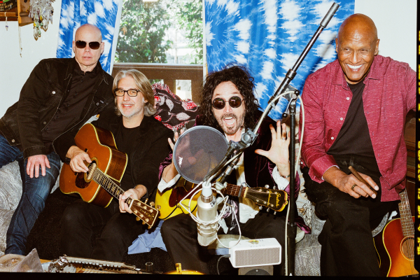 Four men, including Mike Campbell of the Dirty Knobs, sit closely indoors—three hold acoustic guitars near a large mic. Two men smile, one waves with both hands, and one wears sunglasses. A tie-dye blanket and window are visible behind them.