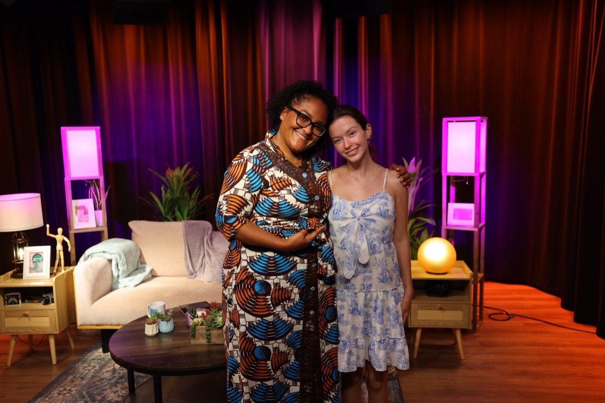 Mikki Wilson, in a long blue, orange, and white print dress, and Ainsley Keating, in a light blue floral sundress, stand side by side with heads touching and smiling in front of a studio set with a beige couch, two pink lamps, plants, and red curtains.
