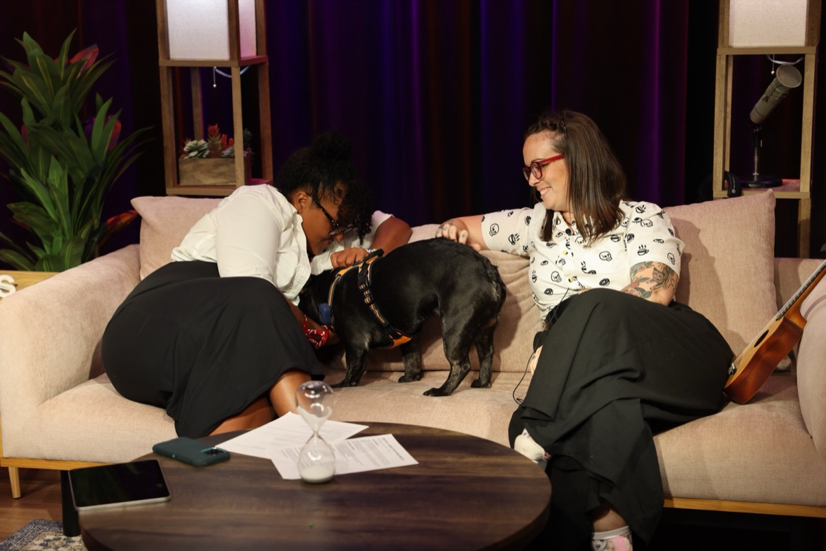 Mikki Wilson and Amanda "A" Larson lean in to pet a black French bulldog standing between them on a beige couch.