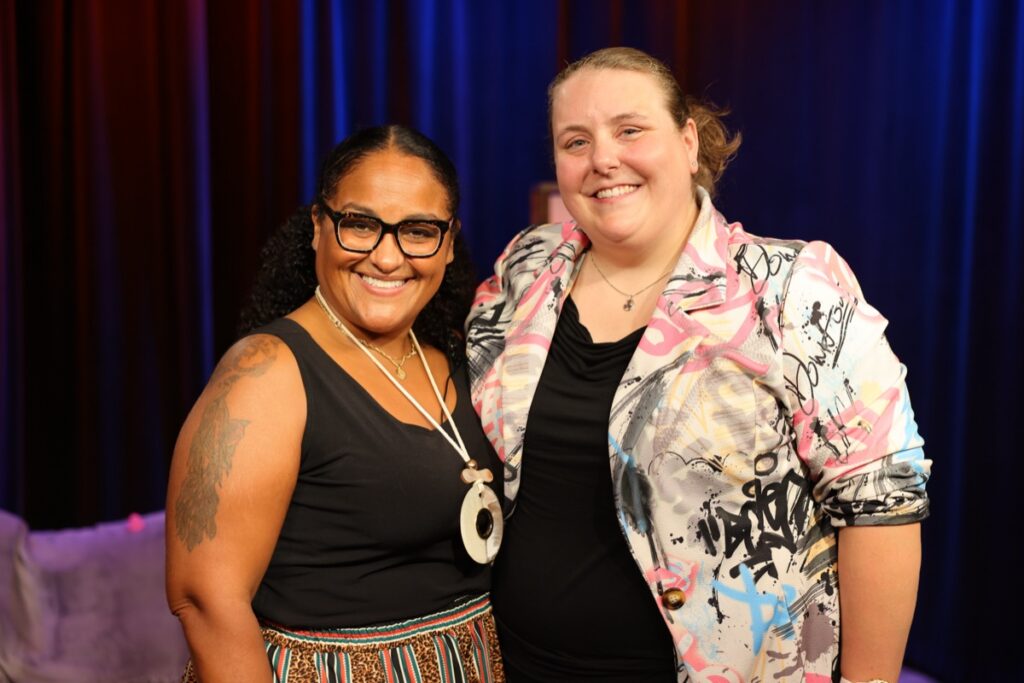 Mikki Wilson, in glasses, a black sleeveless top, and a white pendant necklace, stands shoulder to shoulder with Katie Kimball Dyer, in a white and pink graffiti-print blazer over black, both smiling in front of blue and red studio curtains.