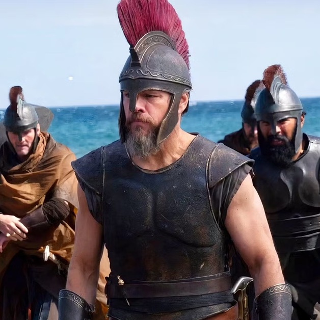 A group of men in ancient-style armor and helmets with red crests stand by the sea; the man in front has a beard, wears a sleeveless breastplate, and looks determined, while others in similar armor are behind him.