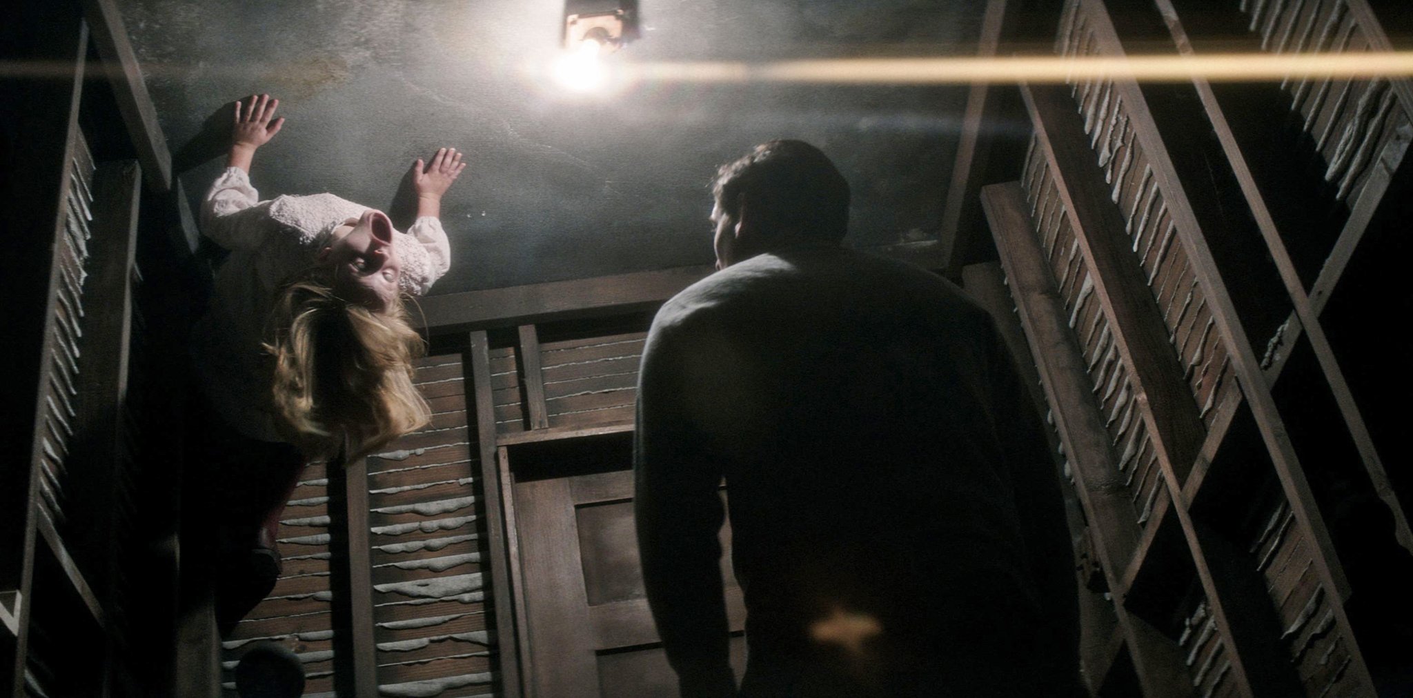 A woman appears to be standing upside-down on the ceiling of a dimly lit, narrow room with exposed wall slats. A man stands on the floor, looking up at her. A single exposed lightbulb hangs from the ceiling, casting dramatic shadows.