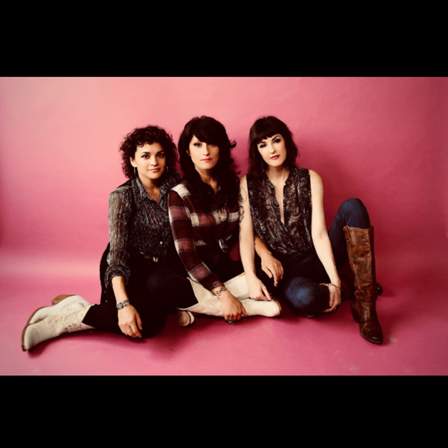 Norah Jones, Catherine Popper, and a third member of Puss N Boots sit on a pink backdrop in boots and jeans, gazing directly at the camera with neutral expressions.