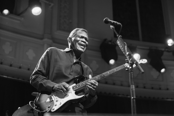 A man stands on stage, smiling while playing an electric guitar. He wears a button-up shirt, performing near a microphone. Stage lights and part of a building interior are visible in the black-and-white image from the Robert Cray Band’s ALL AMPED UP 2026 Tour.