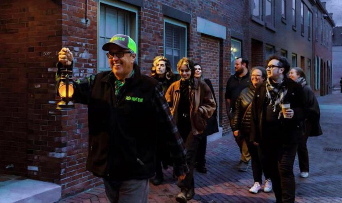 Salem Night Tour founder Tim Maguire smiling and carrying a lantern as he leads a tour group down a brick side street in Salem at dusk