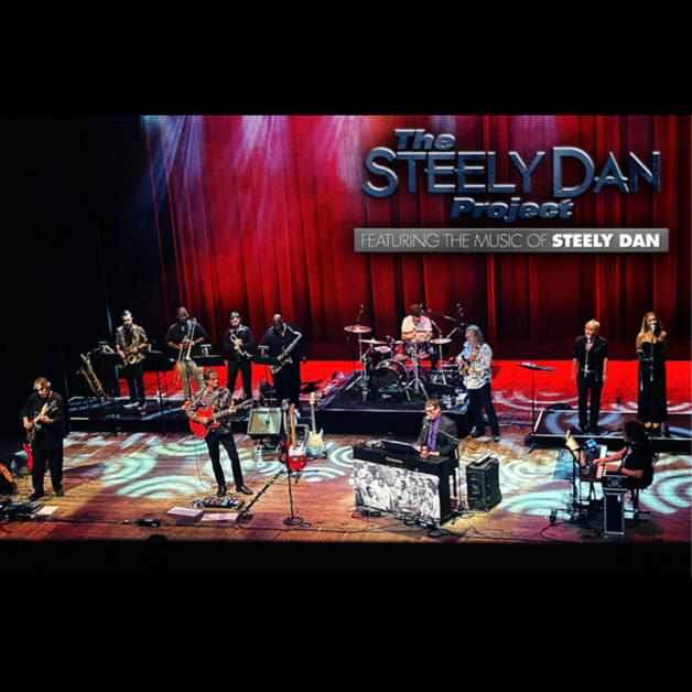 A band of musicians performs on stage with guitars, brass, and keyboards. A sign reads “The Steely Dan Project Featuring the Music of Steely Dan” against a red curtain backdrop as stage lights illuminate their music.