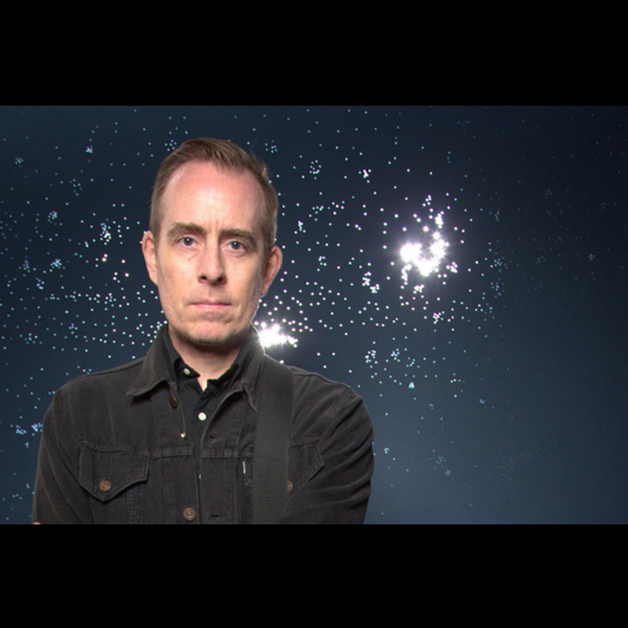 A man with short hair wearing a black jacket stands in front of a backdrop with bright white lights resembling stars or bursts against a dark background. He looks directly at the camera with a neutral expression.