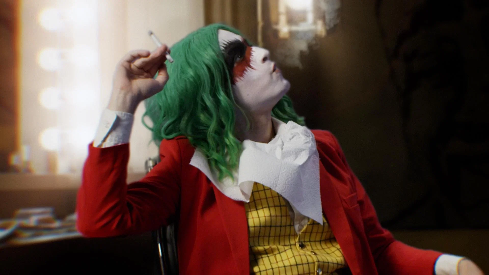 A person with green hair, white face paint, dark eye makeup, and red lipstick wears a red jacket over a yellow plaid shirt and a white bib, holding a cigarette while sitting and looking up. The background is dimly lit, suggesting a dressing room.
