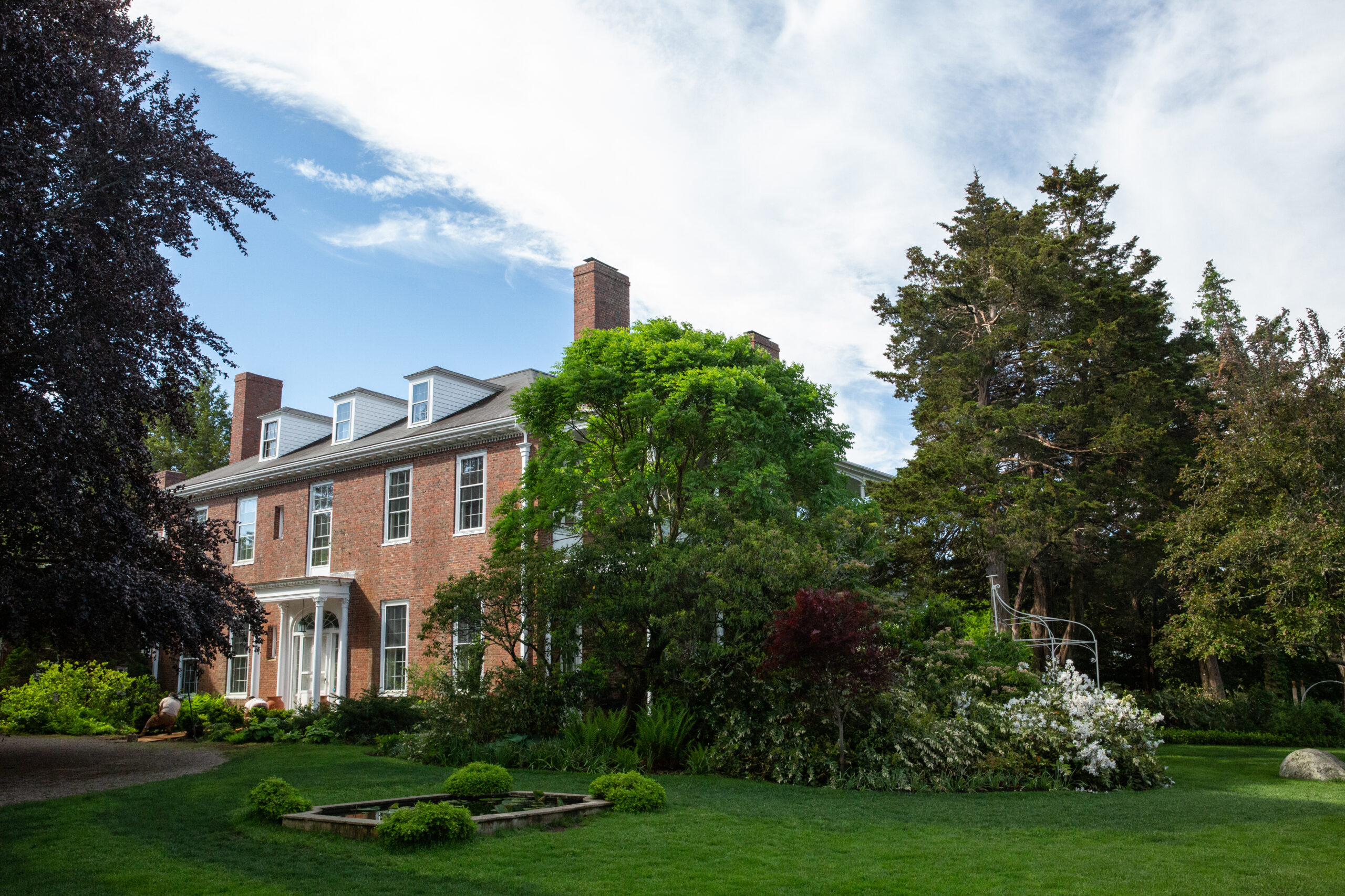 A large brick house with white trim and tall chimneys stands behind a lush green garden and manicured lawn, surrounded by trees under a partly cloudy sky.