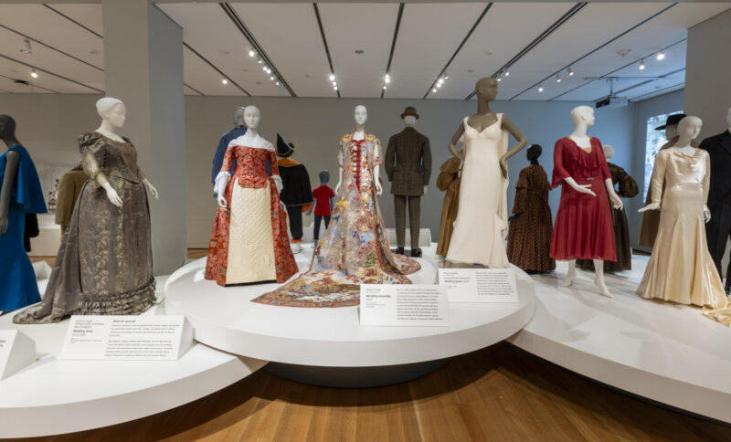 A circular display in a museum gallery featuring several mannequins dressed in various historical and modern wedding gowns and suits, with descriptive placards placed in front of each garment.