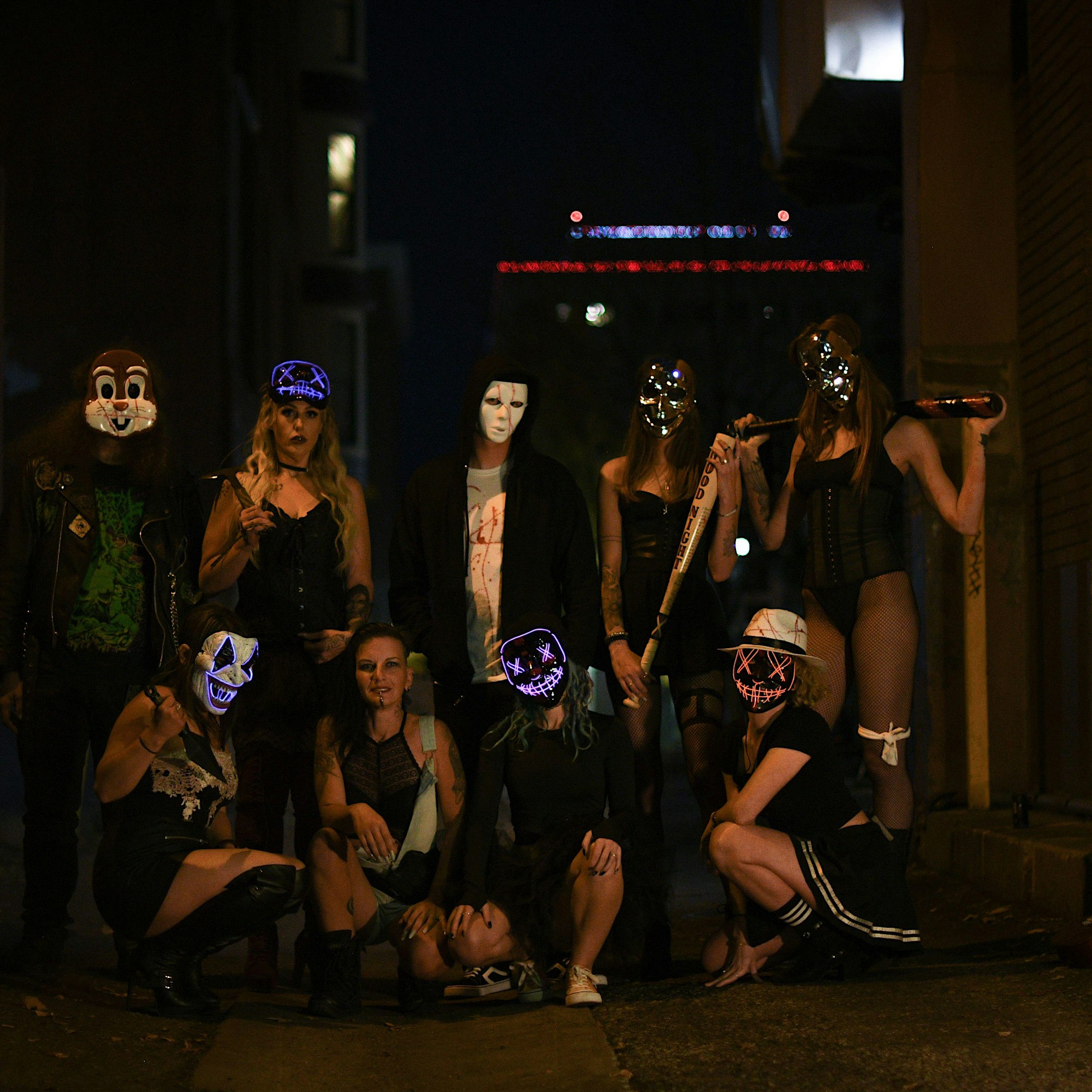 A group of nine people in dark clothing and assorted Halloween masks stand together in an alleyway at night, with some holding baseball bats.