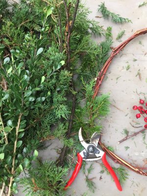 Pruning shears lie on a surface next to evergreen branches, a grapevine wreath base, and a sprig of red berries for a holiday wreath workshop.
