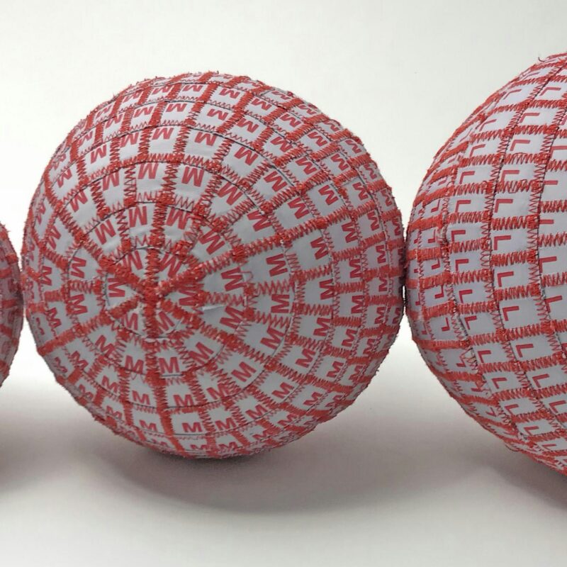 Two spherical objects made of white panels joined by red zig-zag stitching, one patterned with the letter “M” and the other with the letter “L.”