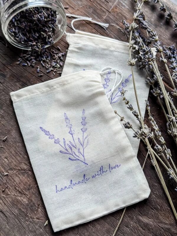 Two muslin pouches with a purple lavender print and the text “handmade with love,” accompanied by dried lavender stems and an open jar of lavender buds on a wooden surface, perfect as natural holiday gifts.