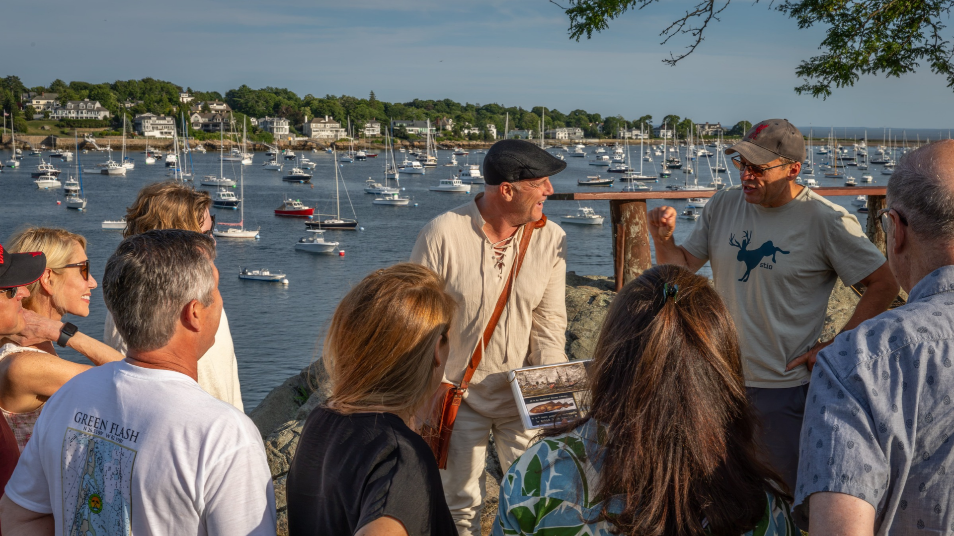 A tour guide in a flat cap speaks to a group of people standing on a rocky overlook above a harbor full of sailboats; this Jay Foss Music tour guide shares the view.