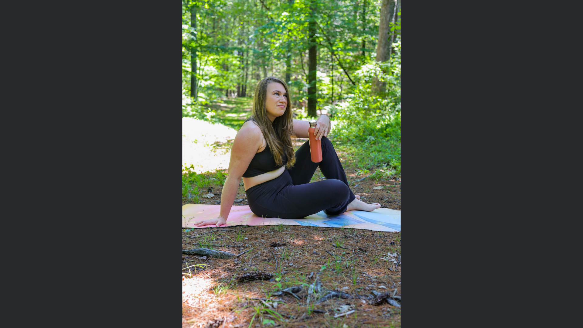 A woman in a black sports bra and leggings sits on a yoga mat in a forest, holding a water bottle, as she meets the member by Lantern Tours.