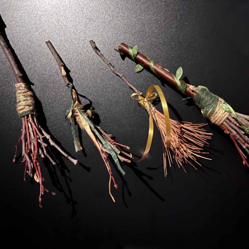 Four miniature brooms made of twigs, twine, and ribbon arranged on a dark surface as part of a weekend workshop.