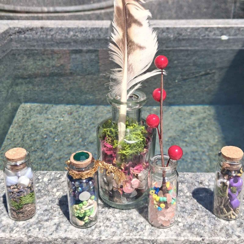 Five small glass magic bottles containing a feather, dried moss, beads, and stones, arranged on a stone ledge as part of a weekend activity in front of the water.