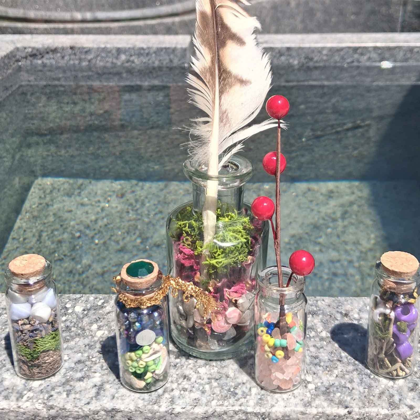 Five small glass magic bottles containing a feather, dried moss, beads, and stones, arranged on a stone ledge as part of a weekend activity in front of the water.