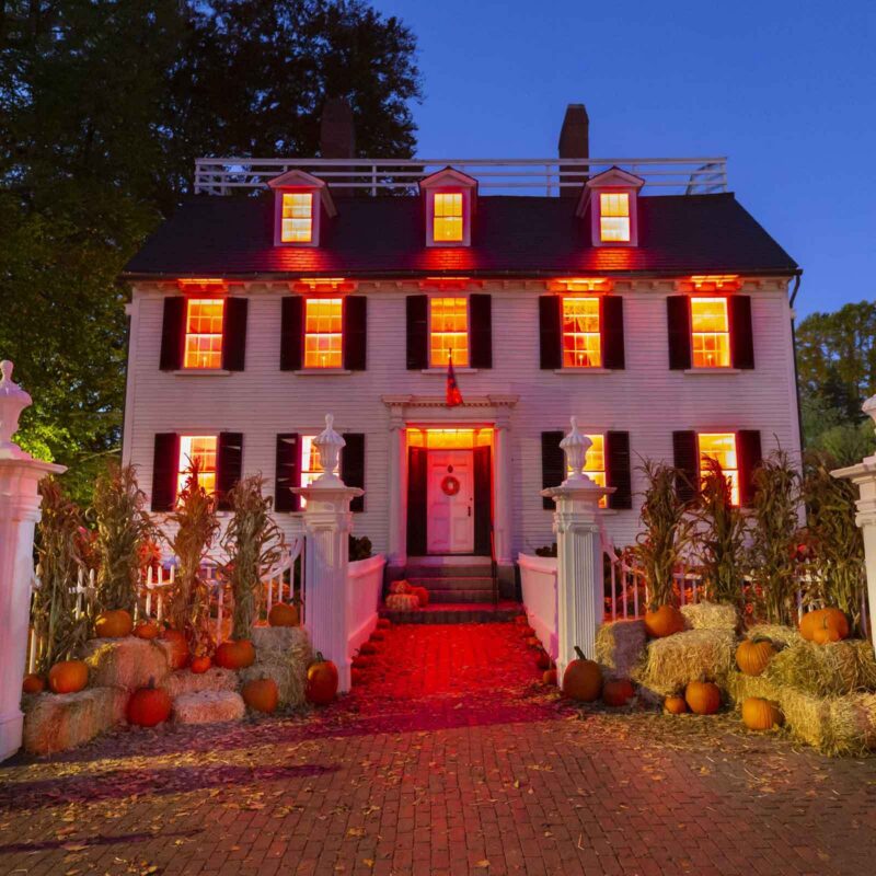 The historic Ropes Mansion decorated for Halloween, with orange-lit windows, stacks of hay bales, and pumpkins lining the brick walkway.