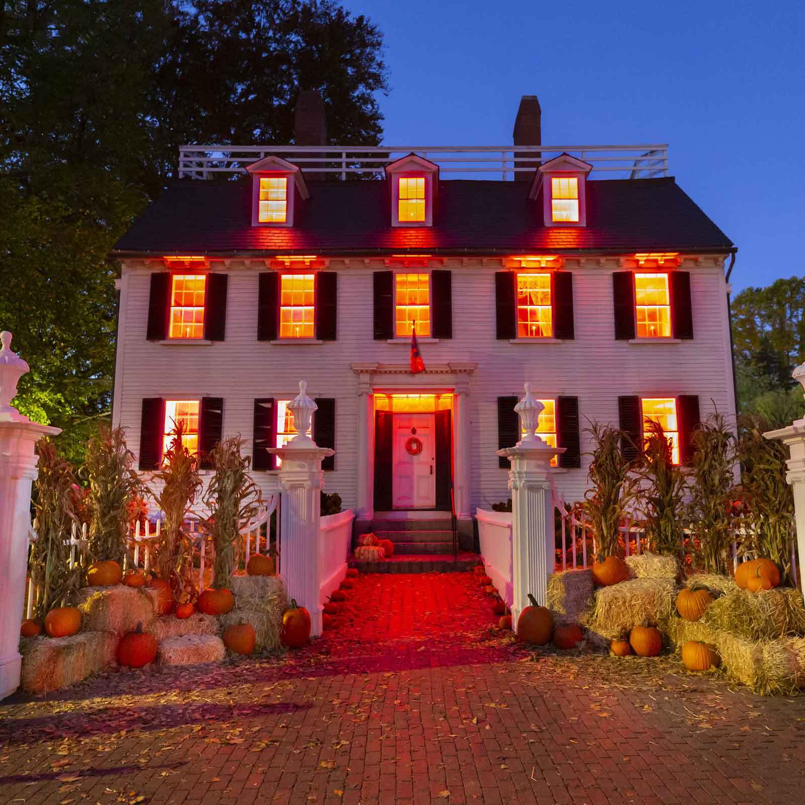The historic Ropes Mansion decorated for Halloween, with orange-lit windows, stacks of hay bales, and pumpkins lining the brick walkway.