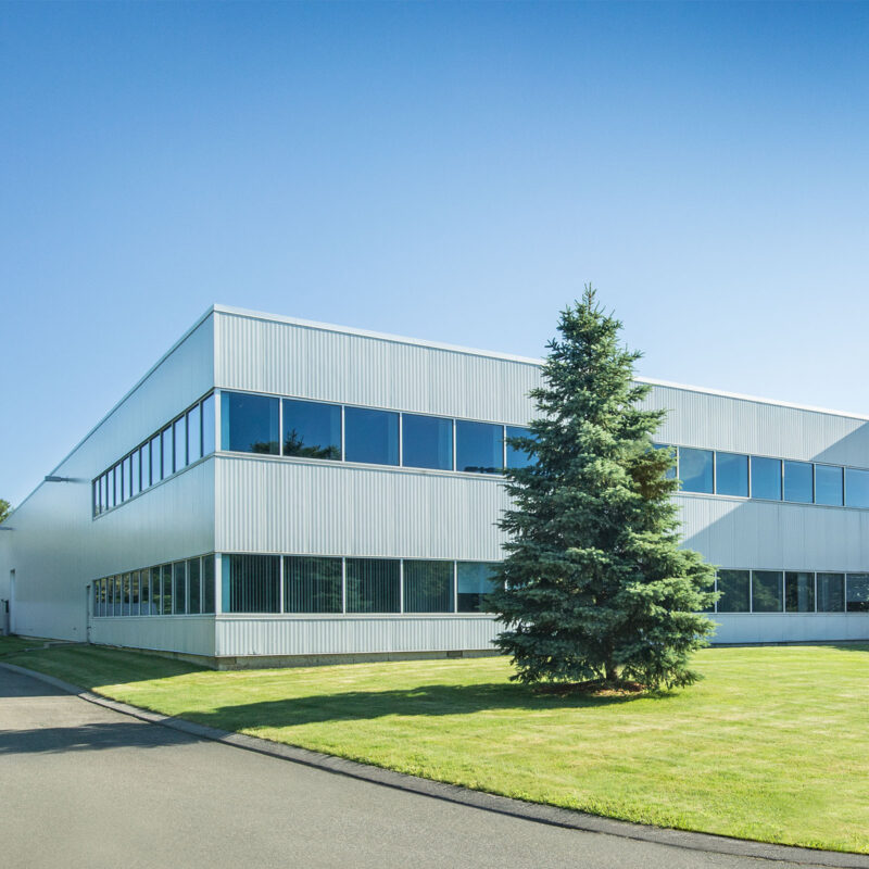 A modern, light-colored industrial building with long rows of windows, situated next to a large pine tree and a grass lawn at the collection center.