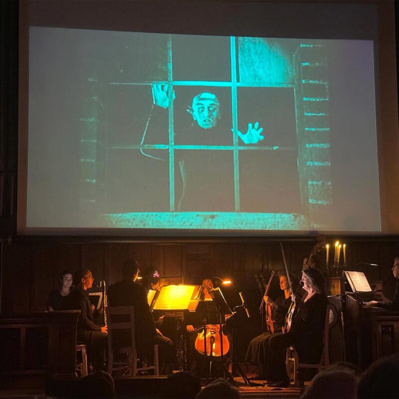 A live orchestra performs on a dimly lit stage in front of a large screen showing a scene from the silent film Nosferatu.