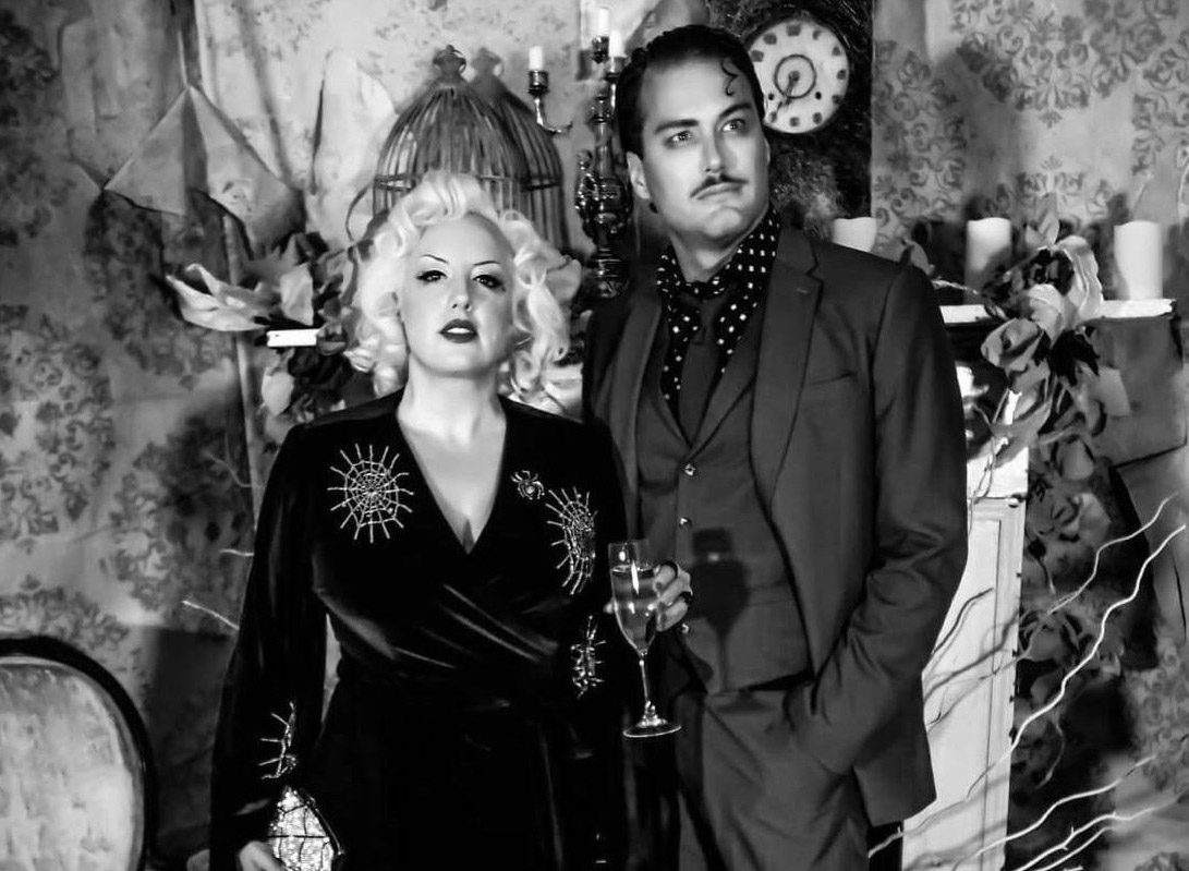 A woman and a man in vintage-style formal attire pose together, with the man holding a glass of champagne.