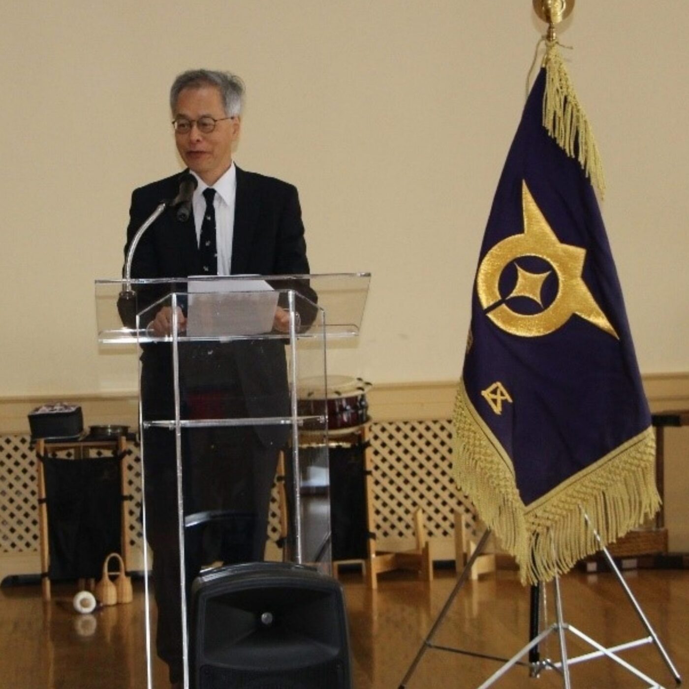 A man in a suit speaks from behind a clear lectern next to a purple and gold flag.