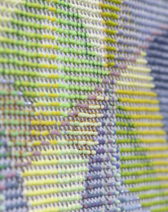 A close-up view of a textured grid pattern, reminiscent of the vessels in contemporary art, made of small, raised beads in shades of yellow, green, and purple.