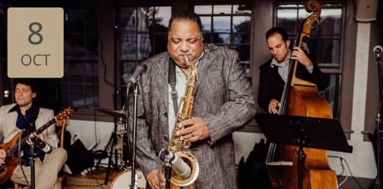A jazz trio performing in a cozy jazz cellar, featuring a saxophonist in the foreground with a guitarist to his left and a double bassist to his right, with a calendar icon reading “8 OCT” overlaid on the top left.