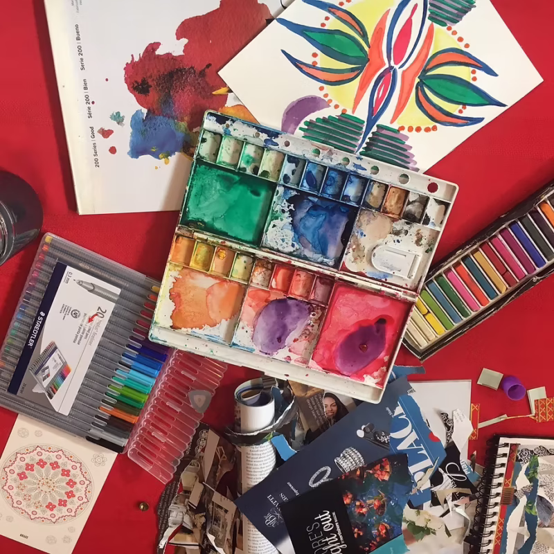 An open watercolor paint set surrounded by fine-liner pens, oil pastels, and paper artworks on a red surface during an art workshop.