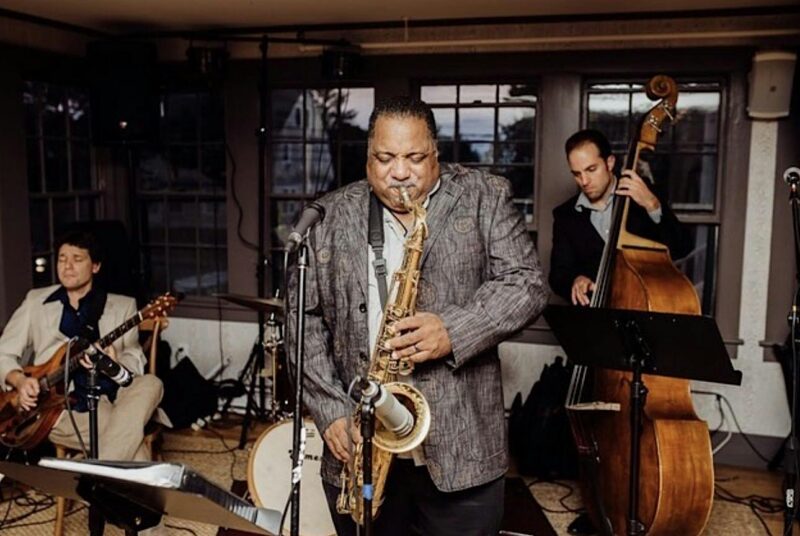 A jazz trio performing indoors, featuring a saxophonist in the center, a bassist on the right, and a guitarist on the left.