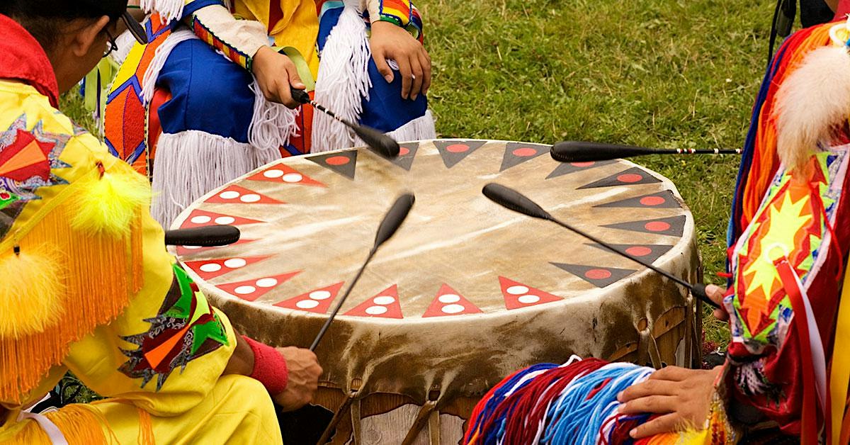 Four people in colorful regalia playing a large circular drum with drumsticks.