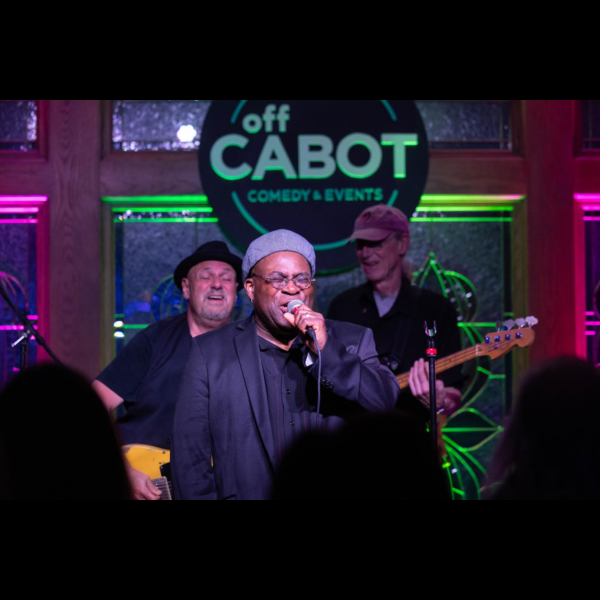 Barrence Whitfield sings into a microphone on stage, accompanied by two musicians playing guitar and bass in front of an Off Cabot Comedy & Events sign.