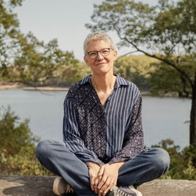Folk singer Catie Curtis, with short grey hair and glasses, sits cross-legged on a rock outdoors with a lake and trees in the background.