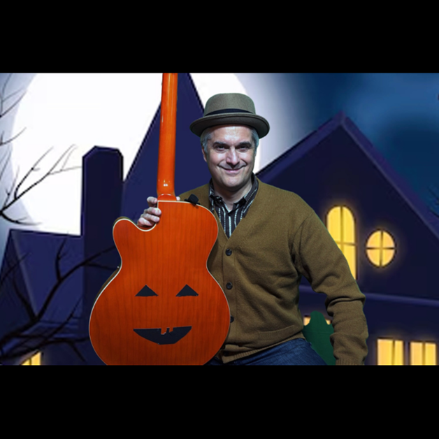 Matt Heaton, wearing a fedora and cardigan, holds an orange jack-o’-lantern acoustic guitar in front of a Halloween-themed backdrop for his Tricks and Treats performance.