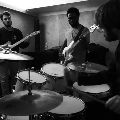 A black and white image of a three-piece band rehearsing in a studio, with a drummer in the foreground, a bassist in the center, and a guitarist on the left.