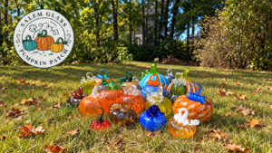 A collection of hand-blown glass pumpkins in various colors scattered on a grassy lawn for Essex County, with a “Salem Glass Pumpkin Patch” logo in the upper left corner.