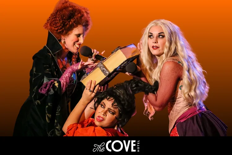 Three performers dressed as the Sanderson sisters from Hocus Pocus hold a book and a microphone during the Rock Cauldron Cabaret, positioned above The Cove Hotel logo.